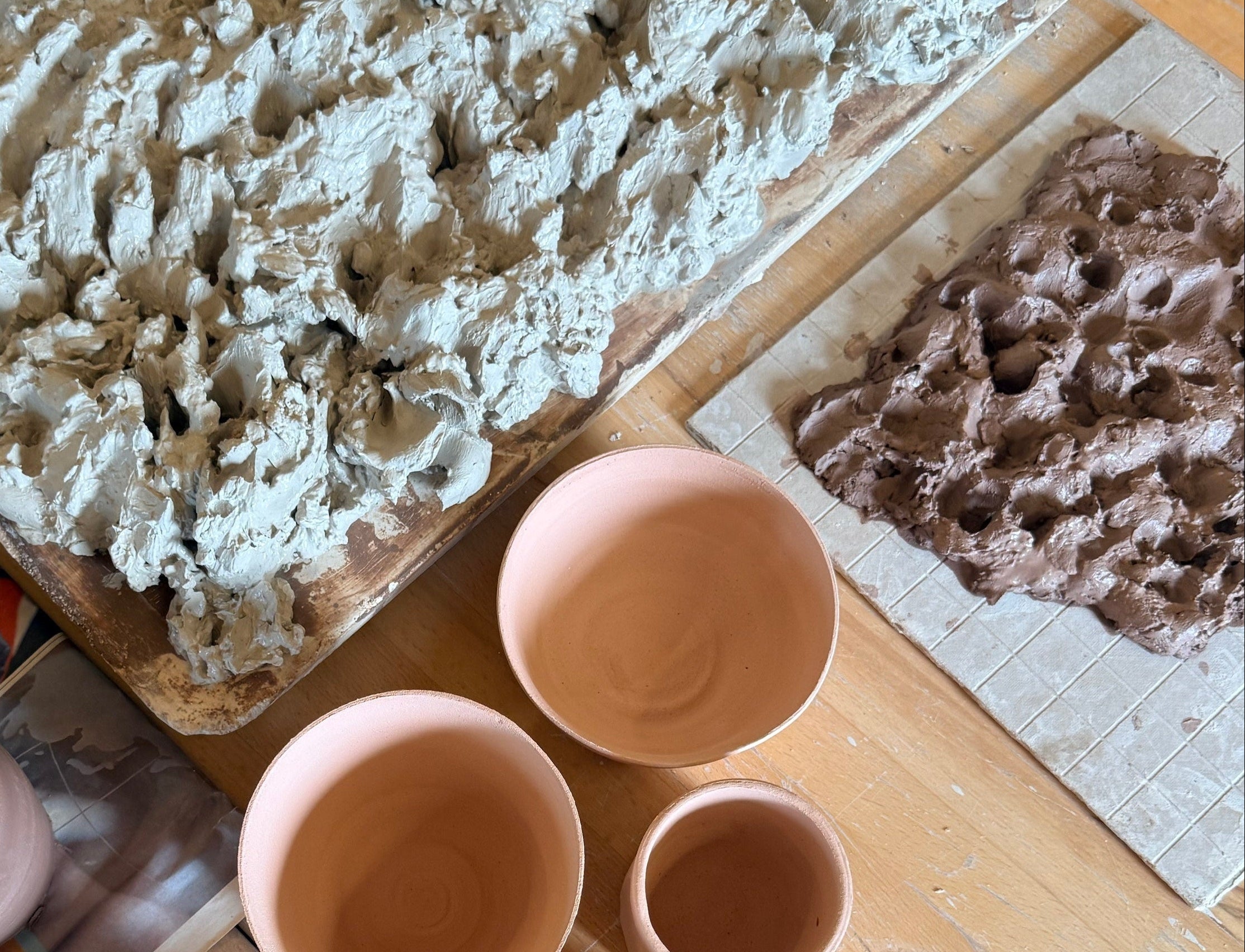 A top-down view of handmade ceramic mugs and trays of clay on a wooden table. The mugs are a dusty terracotta pink, and the trays show textured clay slabs in light gray and brown.