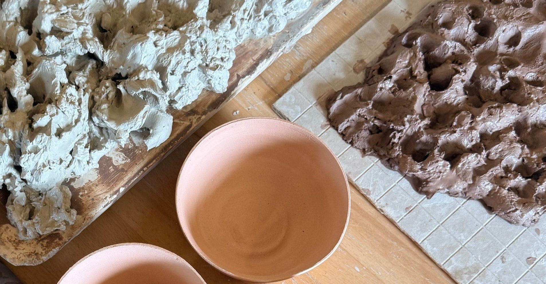 A top-down view of handmade ceramic mugs and trays of clay on a wooden table. The mugs are a dusty terracotta pink, and the trays show textured clay slabs in light gray and brown.