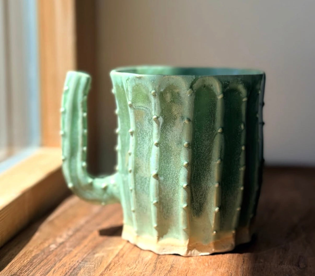 Hand-thrown ceramic mug shaped like a cactus, with a moss green glaze, ribbed body, raised spine details, and a curved arm-shaped handle, sitting on a wooden table near a window.