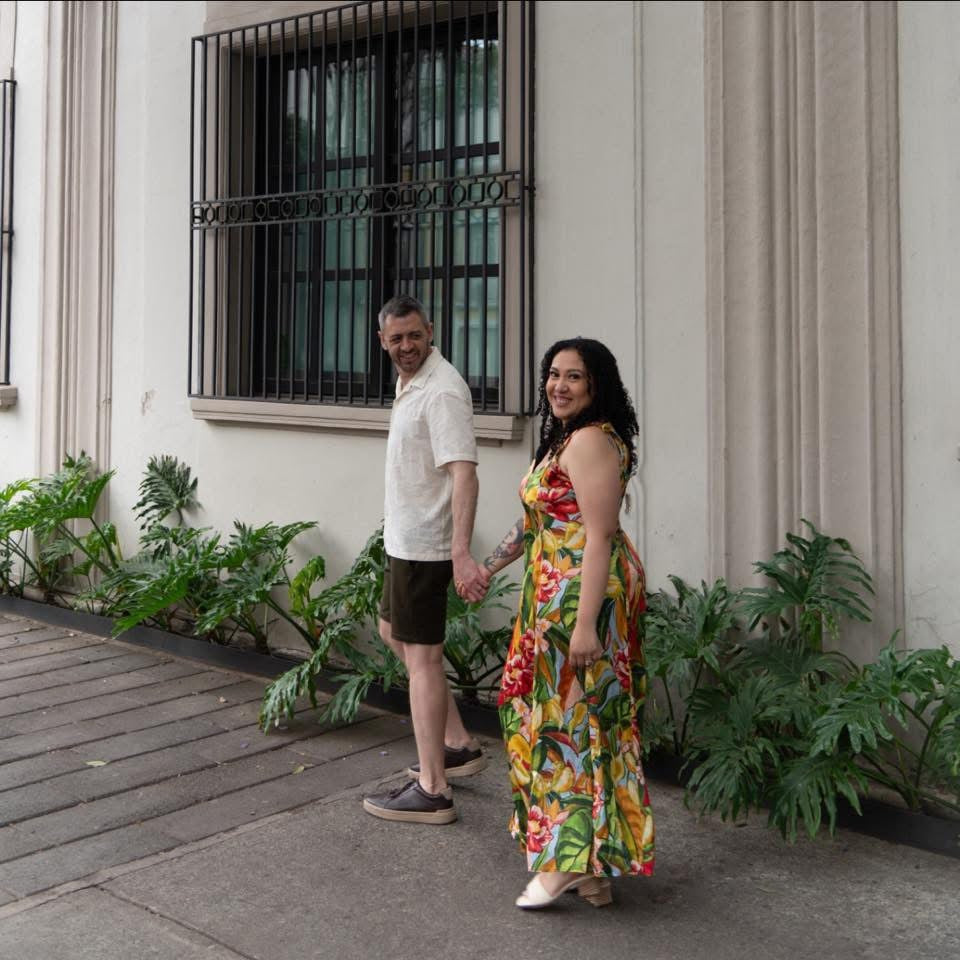 A couple holding hands while walking outdoors on a sidewalk in front of a beige building with a barred window and green plants. The woman is wearing a colorful tropical floral print maxi dress, and the man is wearing a white short-sleeve polo shirt and dark shorts.