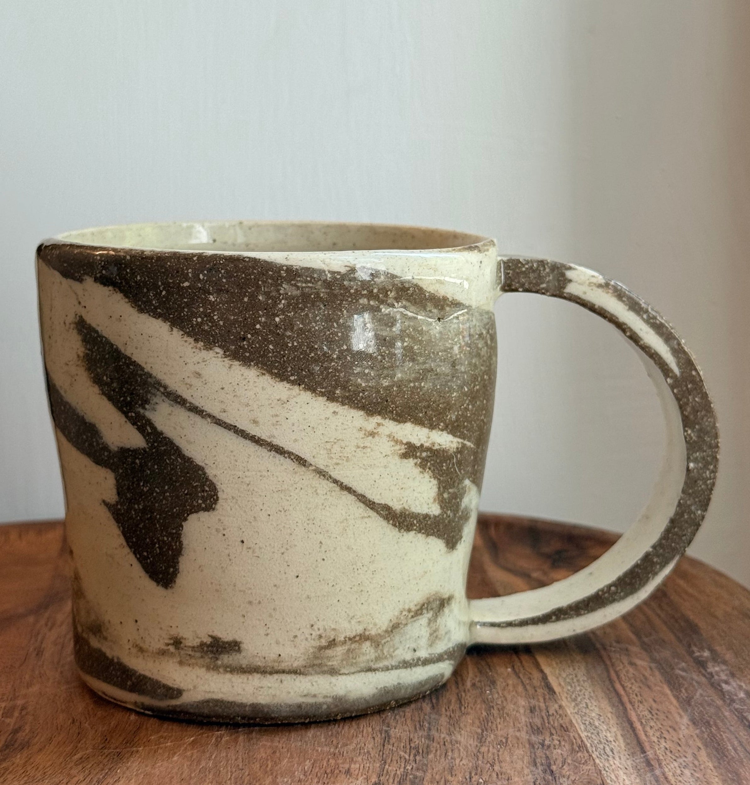 A handmade ceramic mug with a handle, featuring a cream-colored glaze with dark brown and black marbled patterns, shown hanging against a white wall next to a wooden surface.